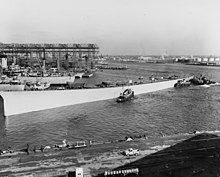 A large hull being pushed through a shipyard by tugboats.